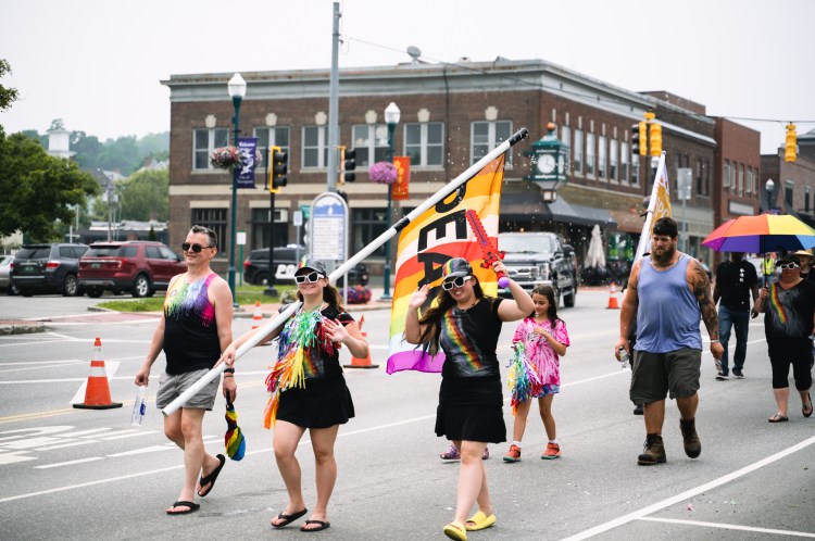 Three people wearing rainbows with large flag that says peace