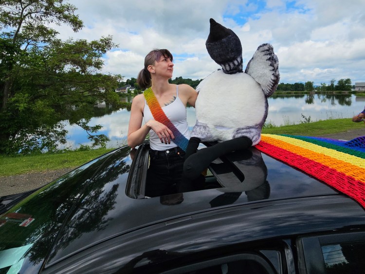 Femme person with large stuffed loon and rainbow sash sticking out of a sunroof